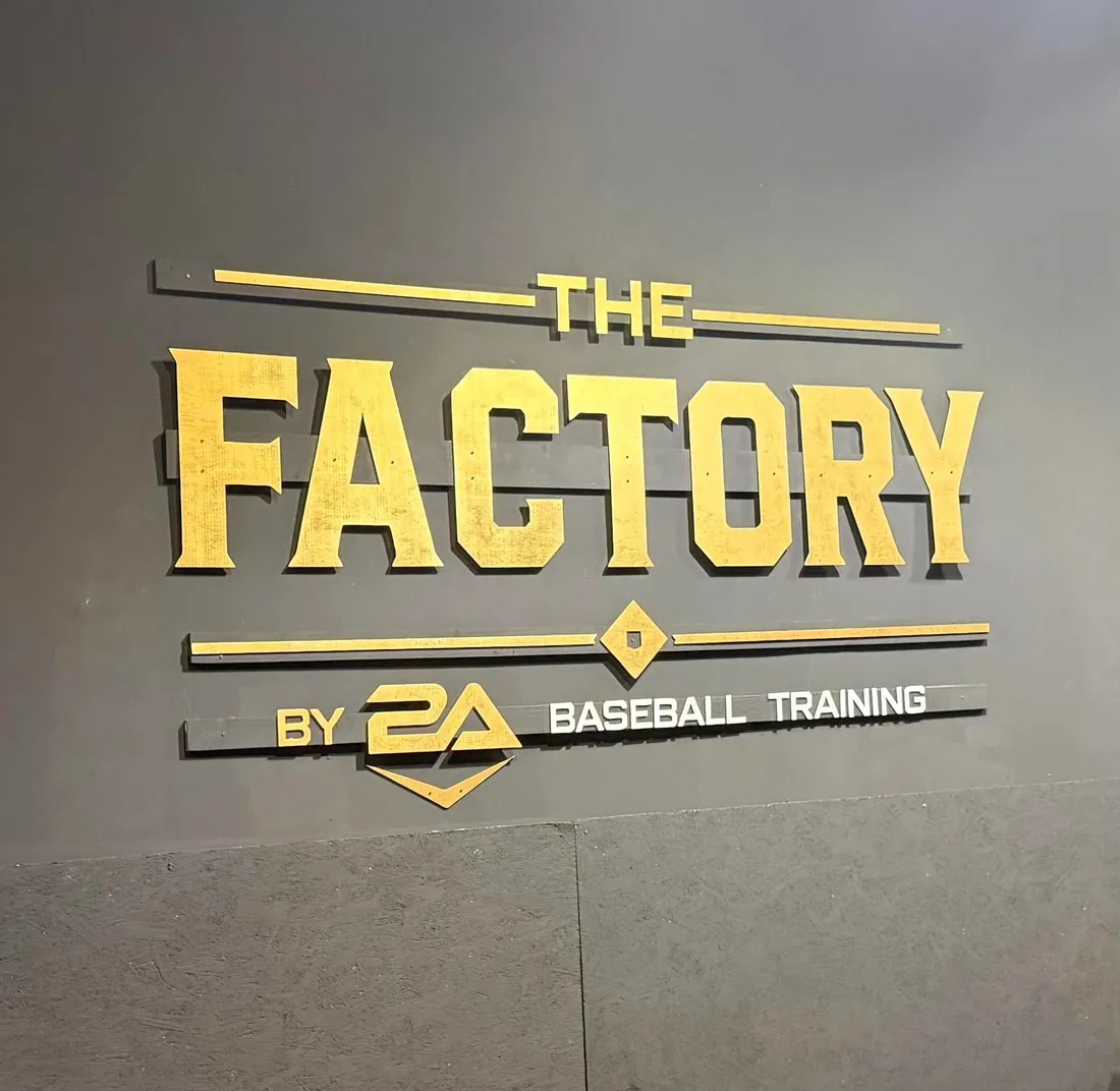 A large routed sign reading The Factory mounted on a wall inside a baseball training facility.