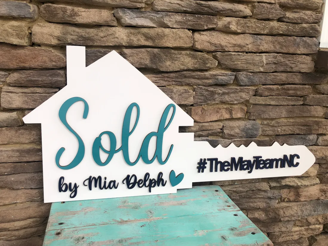 A white house-and-key shaped closing sign reading Sold by Mia Delph in teal script with a hashtag along the key.