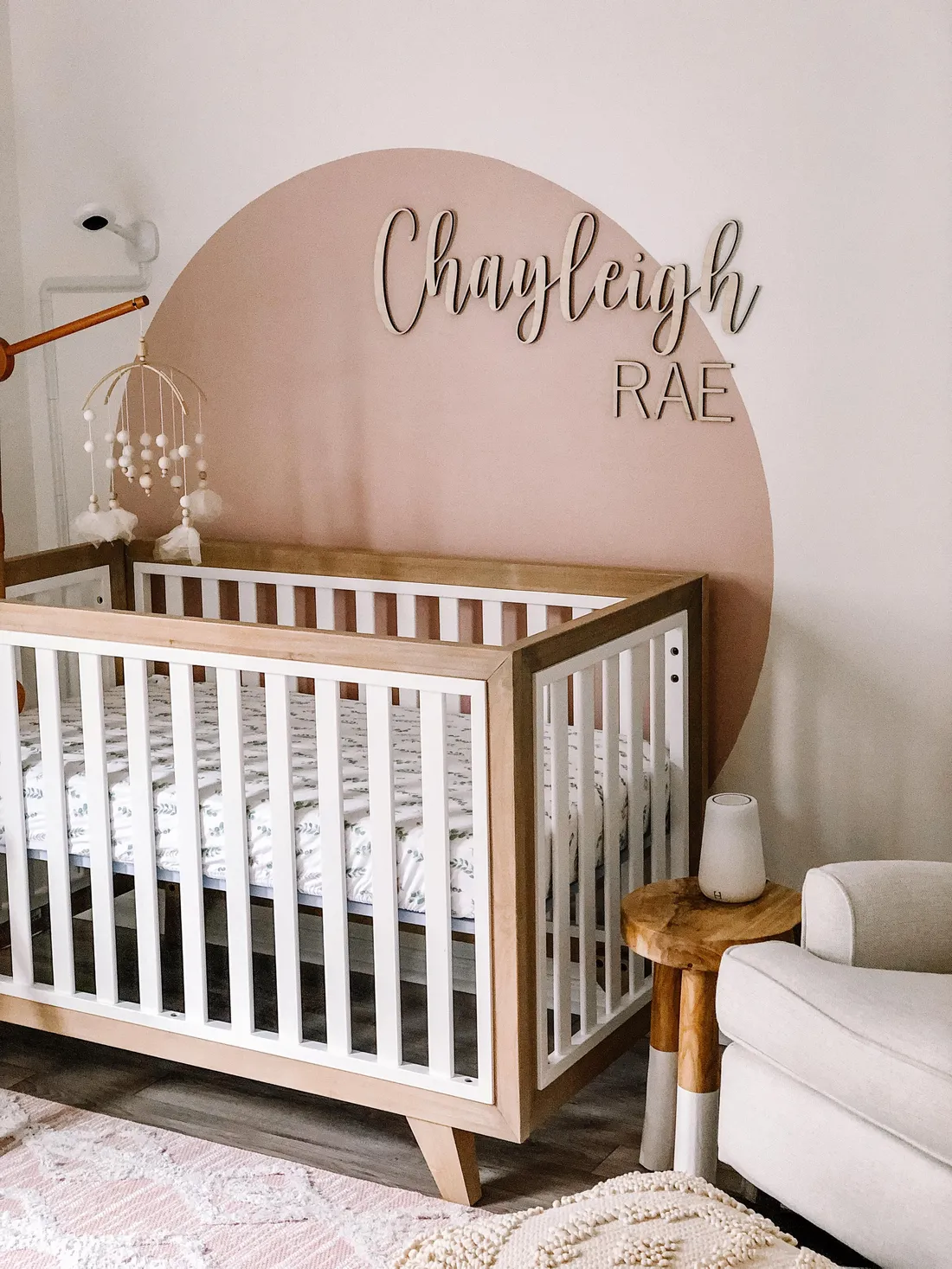 A layered wooden name sign reading Oakleigh Rae above a crib in a pink nursery.
