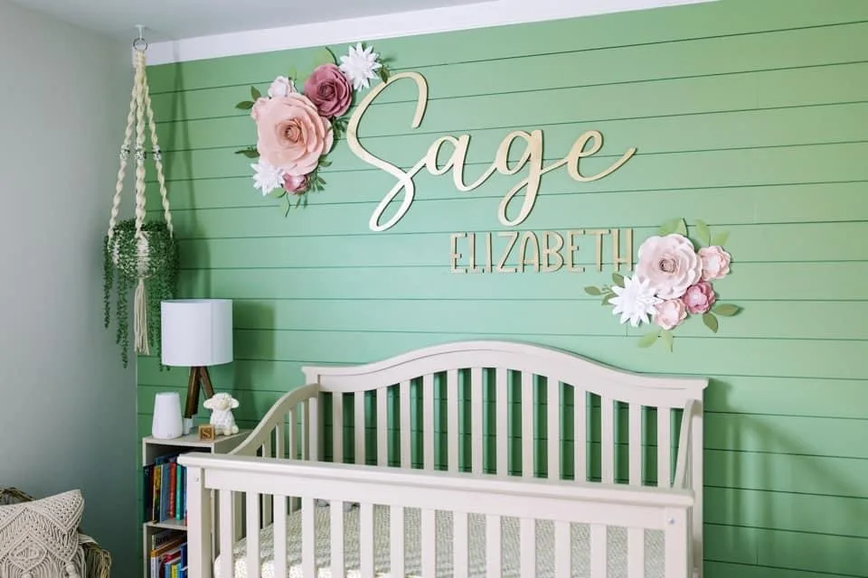 A gold script name sign reading Sage in cursive with Elizabeth beneath it, mounted on a green shiplap nursery wall between two paper flower arrangements, above a white crib.